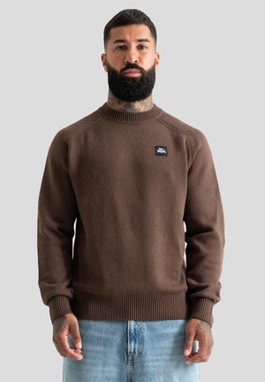 Homme avec barbe et tatouages portant un pull en maille marron et un jean bleu clair, debout devant un fond gris clair.