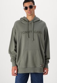 Felpa verde oliva con tasca a marsupio frontale, logo ricamato "TOMMY JEANS" e polsini a costine. Indossata con jeans di denim blu scuro.