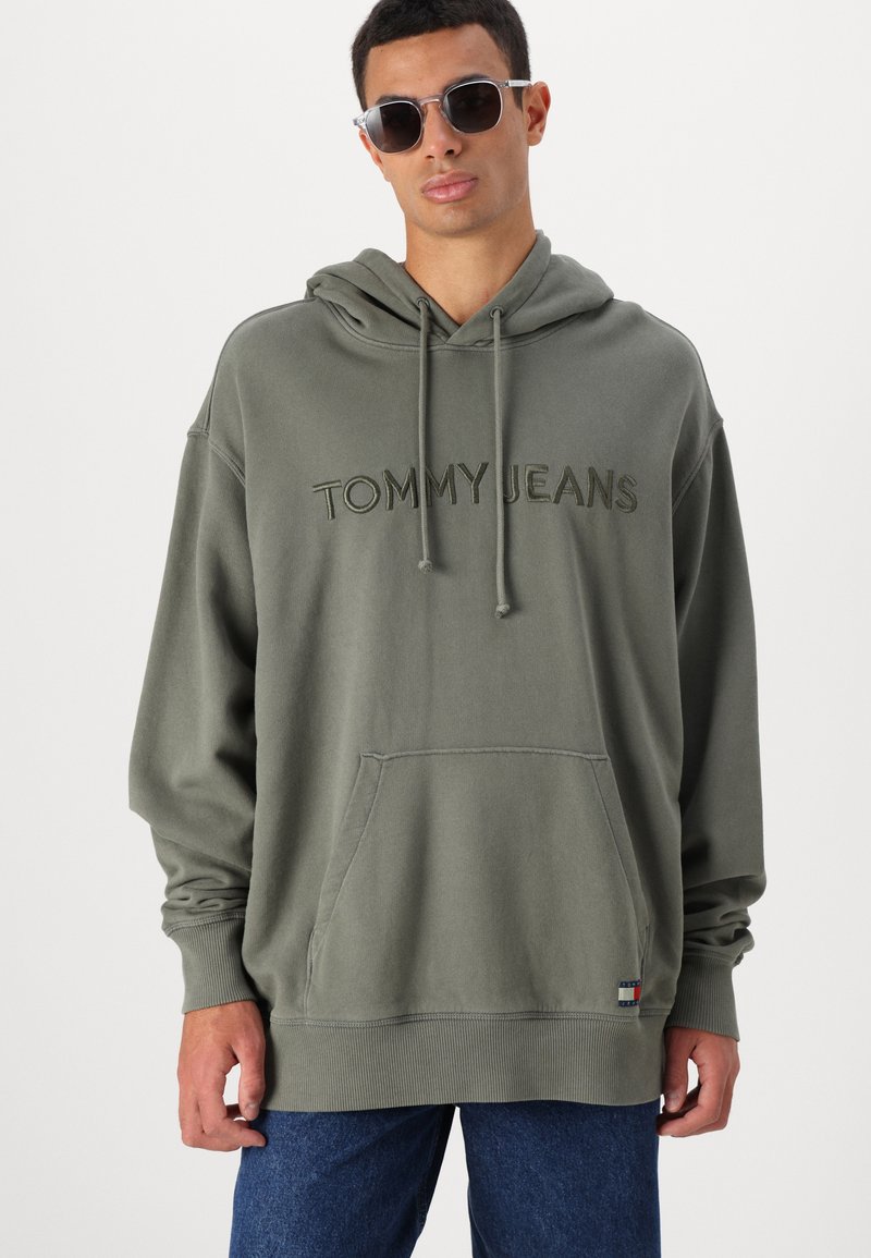 Felpa verde oliva con tasca a marsupio frontale, logo ricamato "TOMMY JEANS" e polsini a costine. Indossata con jeans di denim blu scuro.