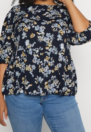 Kvinde iført en mørkeblå blomstret bluse med trekvart lange puffede ærmer kombineret med lyseblå denimjeans.