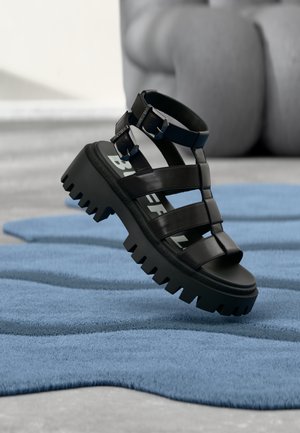 Zwarte chunky sandaal met dikke zool en dubbele gespbanden die boven een blauw gestructureerd tapijt op een lichte vloer zweeft.