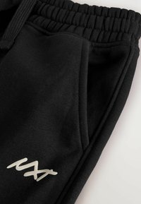 Pantalons de survêtement noirs en tissu doux avec une taille élastique, comportant deux poches latérales et un petit logo blanc sur la cuisse gauche.