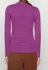 Ribbad lila tröja med långa ärmar och en figurnära form, med en hög halsringning och subtil textur. Paras med bruna byxor.