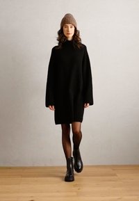 Černé žebrované over size svetr šaty s vysokým límcem a dlouhými rukávy, které jsou doplněny hnědou pletenou čepicí a černými kotníkovými botami. Přírodní dřevěná podlaha.