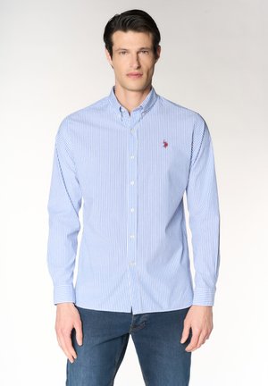 Uomo che indossa una camicia a righe blu e bianca con bottoni e logo rosso ricamato, abbinata a jeans blu, in piedi davanti a uno sfondo uniforme.