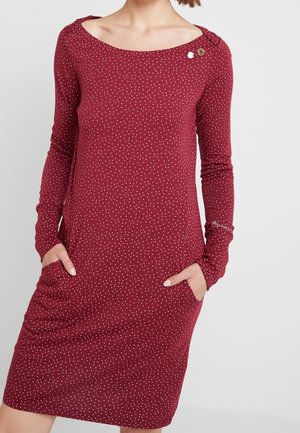 Kobieta ubrana w bordową sukienkę z długim rękawem do kolan, z małymi białymi kropkami i przednimi kieszeniami, ręce w kieszeniach.
