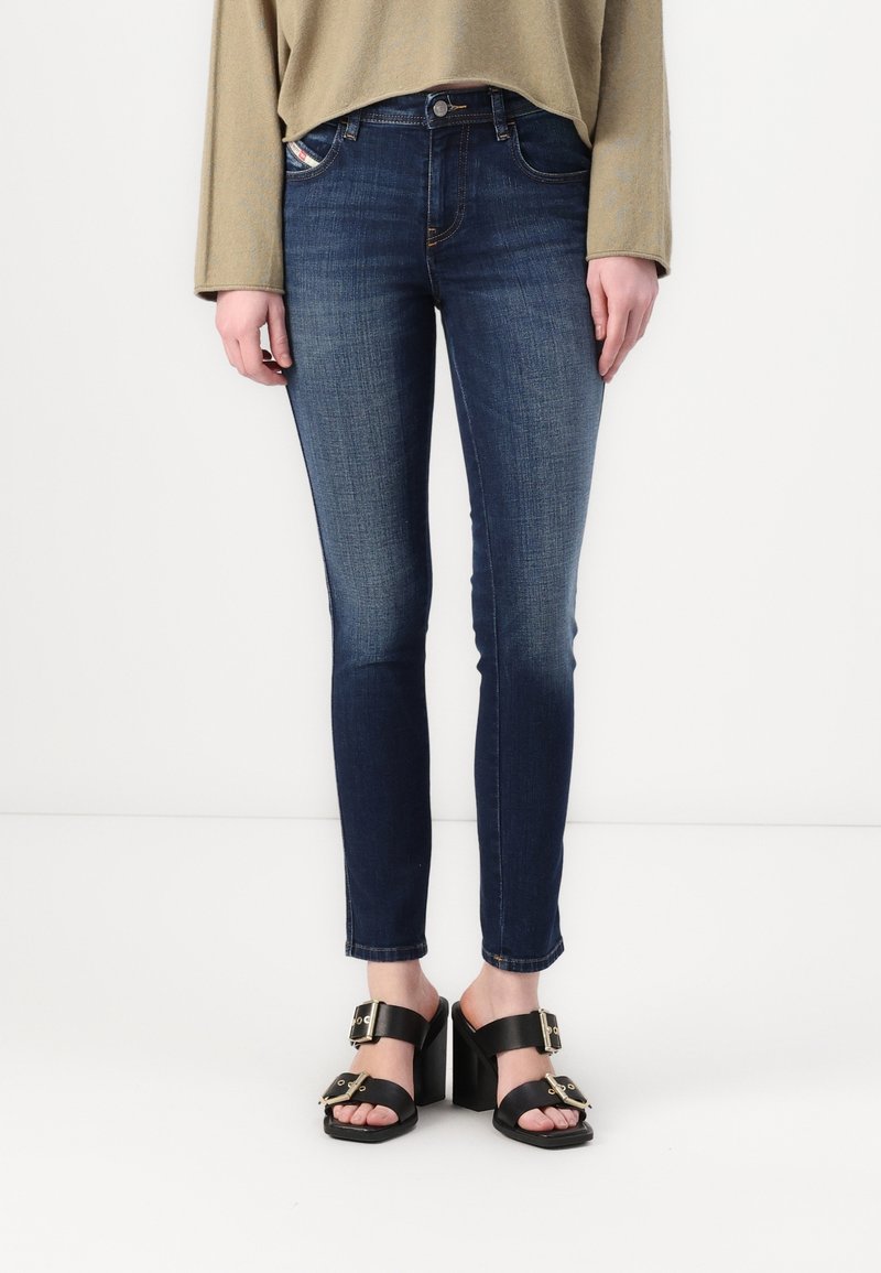 Jeans entallados en denim azul oscuro con una textura suave, que cuentan con bolsillos delanteros y una cintura de altura media, combinados con sandalias de tacón negras.
