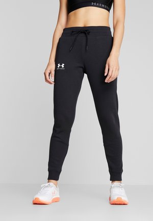 Vrouw draagt zwarte Under Armour joggingbroek, zwarte sportbeha en witte sneakers met oranje zolen, staand tegen een effen grijze achtergrond.