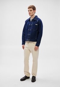 Denimjakke i mørk blå med knapper foran, kontrasterende hvit søm, brystlomme og krage; kombinert med beige bukser og svarte sko.