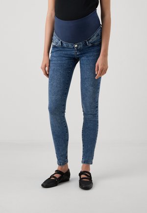 Pregnant person wearing blue maternity jeans with a high elastic waistband and black flat shoes with straps.