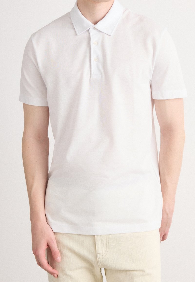 Homme portant un polo blanc uni à manches courtes et un pantalon de couleur claire, debout avec une main détendue le long du corps.