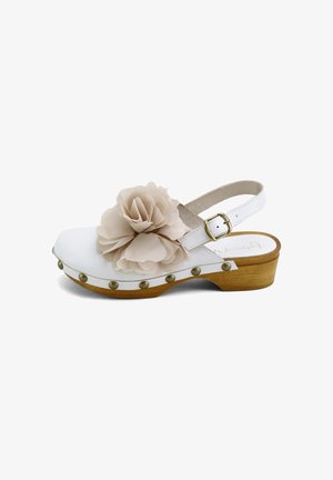 Sandalia tipo zueco de cuero blanco con suela de madera, tachuelas de latón, correa en el tobillo y una gran flor de tela beige en la parte superior.