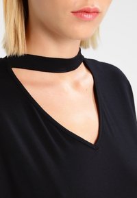 Top negro con cuello en V y un distintivo collar estilo choker, fabricado en un tejido suave con una textura delicada.