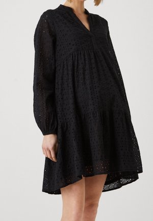 Vrouw draagt een losse, zwart eyelet-jurk met lange mouwen, een V-hals en een roklengte boven de knieën met laagjes.