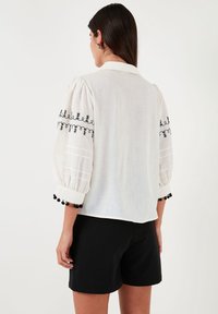 Blouse blanche avec des manches bouffantes, ornée de broderies noires et de pompons aux poignets. Associée à un short noir.