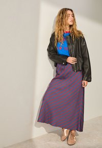 Giacca di pelle nera, t-shirt grafica blu con testo rosso, gonna maxi a righe rosse e blu, sneakers marroni, capelli mossi, sfondo chiaro neutro.