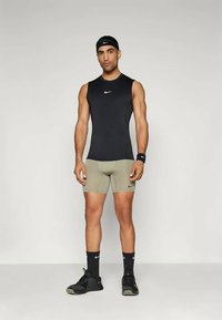 Athletischer Mann steht, trägt ein schwarzes ärmelloses Oberteil, khakifarbene Kompressionsshorts, ein schwarzes Stirnband, ein Armband, Socken und Sneaker, auf einem schlichten Hintergrund.