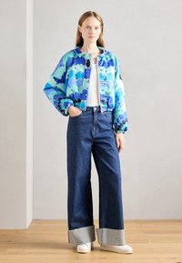Giacca bomber trapuntata in un motivo floreale blu e verde, abbinata a jeans a vita alta e gamba larga con orli a contrasto e sneakers bianche.