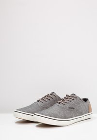 Zapatillas de tela gris con acentos en el talón marrón, suela de goma blanca y diseño de cordones. Textura suave, forma redondeada y detalles de costura en contraste.