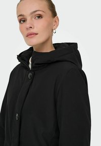 Crna kapuljača jakna sa patentnim zatvaračem, s dugmadima kao naglaskom i glačkom teksturom. Ima visoki ovratnik i uski kroj.