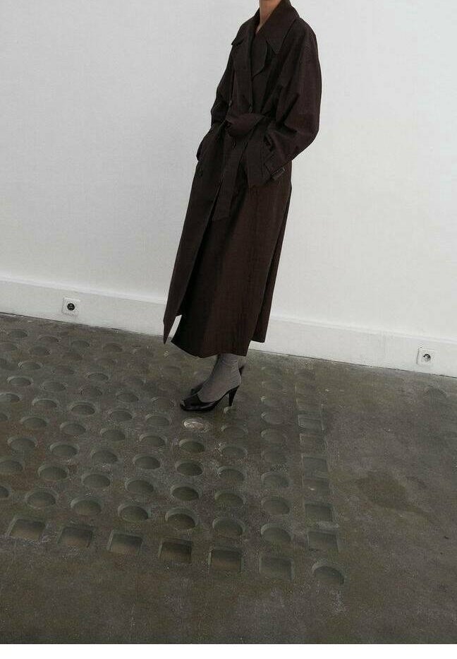Personne portant un long trench-coat sombre, des chaussettes grises et des chaussures à talons noirs, debout sur un sol gris texturé.