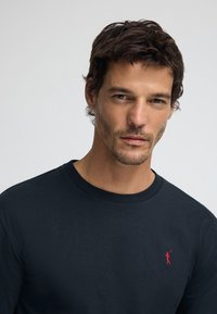 Svart bomullslångärmad tröja med rund halsringning, med en liten röd broderad logotyp på vänster bröst. Mjuk textur.