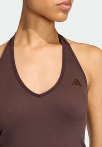 Hnědý sportovní top s výstřihem do V a tenkými ramínky. Má texturovanou látku a nenápadný logo akcent na levé straně.