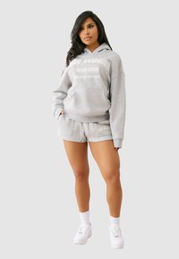 Szara bawełniana bluza z kapturem z przednią kieszenią i nadrukiem logo, w połączeniu z odpowiednimi szarymi szortami. Posiada ściągacze na rękawach i białe sneakersy.