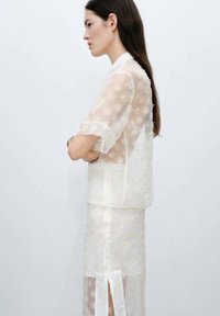 Top trasparente ricamato con fiori e maniche corte; vestito a strati sottostante con uno spacco all'orlo; colori morbidi bianco e giallo.