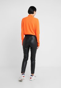 Maglione arancione a maniche lunghe con collo alto, abbinato a pantaloni in pelle nera aderenti e sneakers bianche con dettagli rossi e blu.