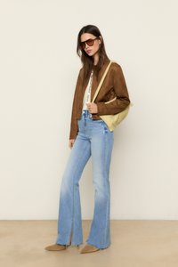 Veste en daim marron, top blanc, jeans évasés bleus clairs avec des fentes sur le côté, et ballerines pointues marron. Accessoirisée avec un sac à bandoulière jaune et des lunettes de soleil.