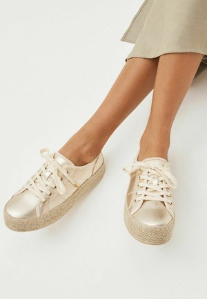 Jambes portant une jupe beige et des baskets compensées dorées métalliques à lacets avec semelles en corde tressée sur fond blanc.