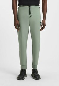Lichtgroene joggers van glad materiaal, met een elastische tailleband en een trekkoord, en taps toelopende boorden. Zwarte hardware-accenten zichtbaar.
