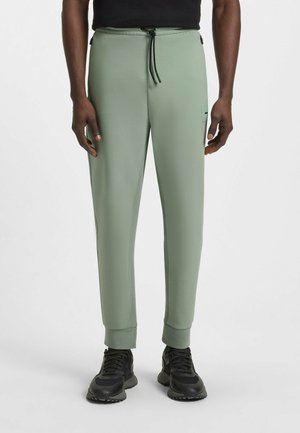 Ljusgröna joggare i mjukt material, med ett elastiskt midjeband och snörning, samt smalnande ärmslut. Svarta metalldetaljer synliga.