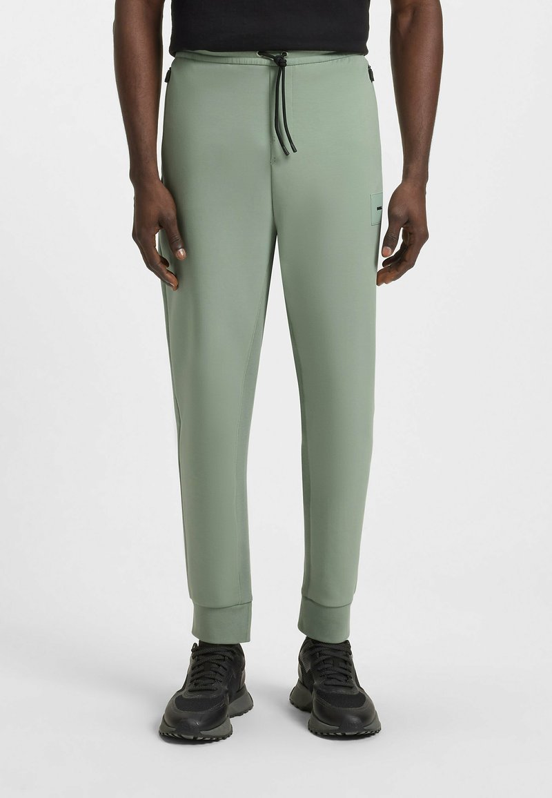 Lichtgroene joggers van glad materiaal, met een elastische tailleband en een trekkoord, en taps toelopende boorden. Zwarte hardware-accenten zichtbaar.