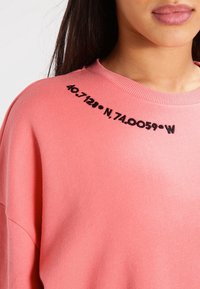 Sudadera rosa con coordenadas negras bordadas en el hombro. Tejido suave con un corte holgado y cuello acanalado.