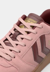 Scarpa sportiva in suede rosa con lacci crema, accenti bordeaux e design a chevron. Include una linguetta con logo dorato sulla linguetta e una suola testurizzata.