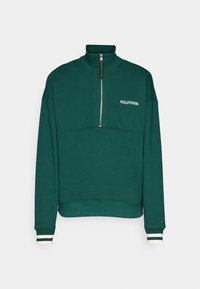 Sudadera verde con cuello alto, media cremallera y logo blanco. Puños acanalados con rayas verdes y blancas. Tejido suave y texturizado.