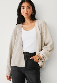 Cardigan beige avec une texture douce et une ouverture sur le devant. Comprend des manches larges et est porté sur un débardeur blanc et un pantalon noir.
