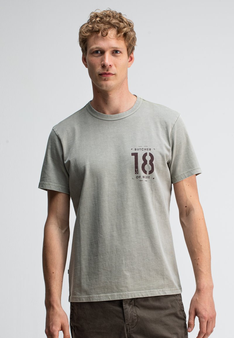 Hellgraues Baumwoll-T-Shirt mit Rundhalsausschnitt, kurzen Ärmeln und einem dunklen Druck mit den Zahlen "18" und dem Wort "BUTCHER" auf der Vorderseite.