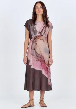 Nő ujjatlan midi ruhában, amelynek mintája absztrakt rózsaszín, barna és bézs árnyalatú, folyékony hatású, karjai lazán a test mellett, és világosbarna szandált visel.