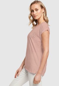 Lyserød bomulds-T-shirt med korte ærmer og rund halsudskæring. Har et enkelt sort hjerte- og kløverdesign på forsiden.