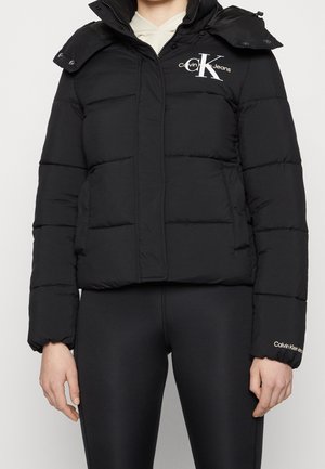 Czarna puchowa kurtka z logo Calvin Klein Jeans na piersi i rękawie, noszona na kremowej bluzie z kapturem, zestawiona z czarnymi legginsami.