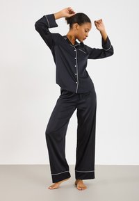 Svart sateng pysjsett med en knappdown skjorte og vide bukser, med hvit piping langs kragen, mansjettene og nederkanten.