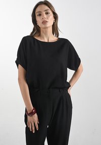 Čierny krátky rukáv top z ľahkej látky s okrúhlym výstrihom, skombinovaný s čiernymi nohavicami s vysokým pásom a červeným náramkom na zápästí.
