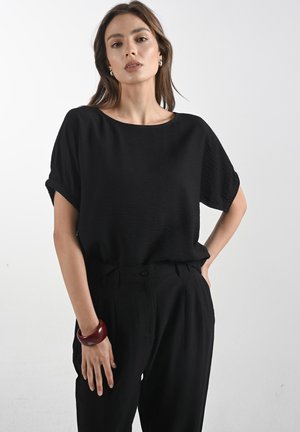 Černý top s krátkým rukávem vyrobený z lehkého materiálu s kulatým výstřihem, kombinovaný s černými kalhotami s vysokým pasem a červeným náramkem na zápěstí.