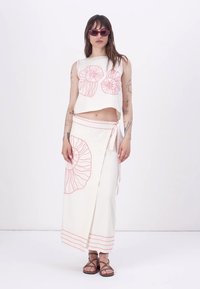 Top corto bianco con motivi a conchiglia rosa, abbinato a una gonna bianca con dettagli coordinati e un fiocco in vita. Sandali marroni.