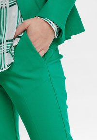 Zielone dopasowane spodnie o gładkiej fakturze i dopasowanym kroju. Na zdjęciu widoczna jest ręka w bocznej kieszeni, a pod spodem widać wzorzystą koszulę.