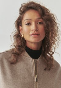 Světle béžová bomber bunda vyrobená z vlněné směsi, s ribbovaným límcem a zlatým zipem. Model má vlnité vlasy a nosí odvážné náušnice.