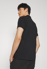 Man in black short-sleeve polo shirt and black pants, standing with hands in pockets, looking over shoulder against plain background.